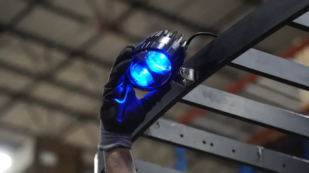A gloved hand installs a blue-glowing dual-lens LED industrial light on a metal beam in a warehouse.