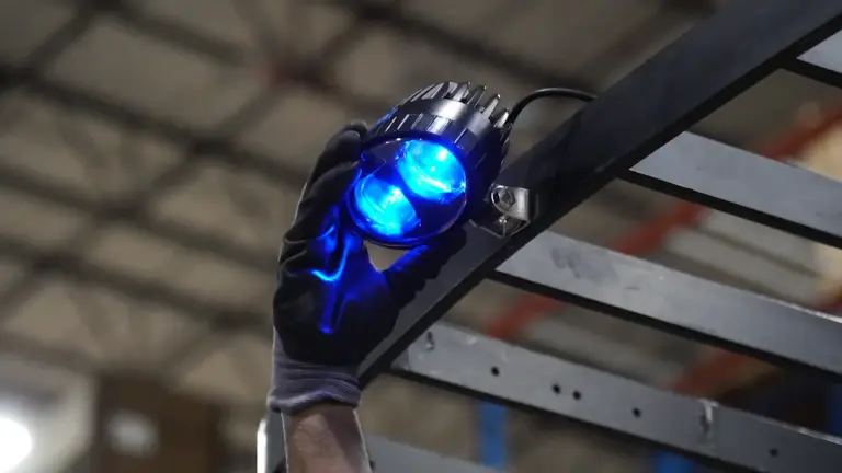 A gloved hand installs a blue-glowing dual-lens LED industrial light on a metal beam in a warehouse.
