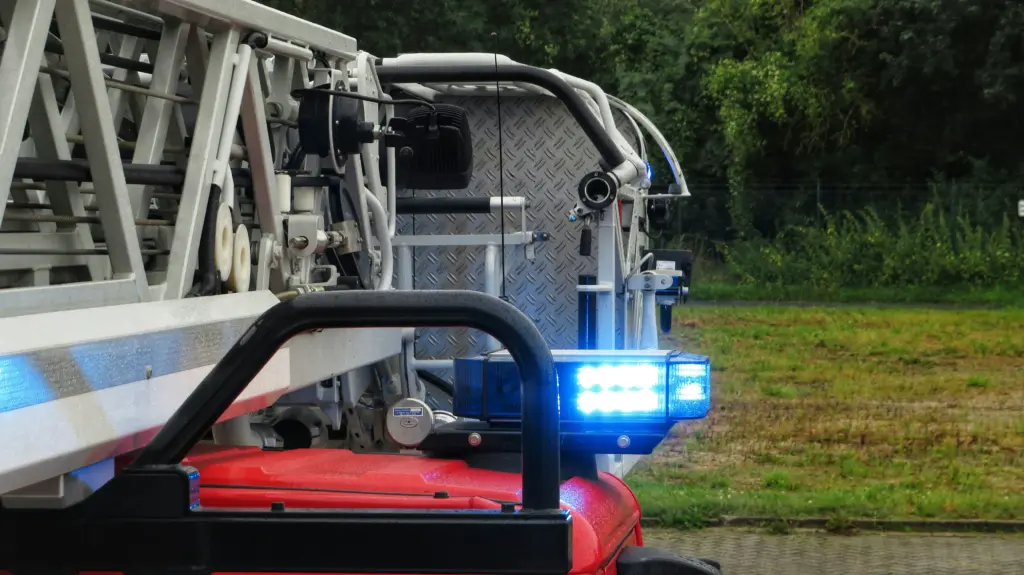 Side view of a fire engine showing its ladder, hoses, and bright blue flashing emergency lights parked on grass.