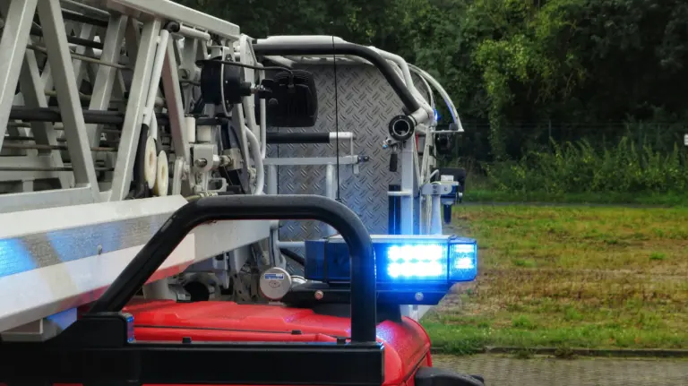 Side view of a fire engine showing its ladder, hoses, and bright blue flashing emergency lights parked on grass.