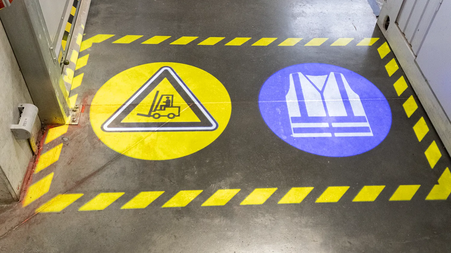 Warehouse floor with painted hazard markings and safety signage that a laser line projector augments in real time